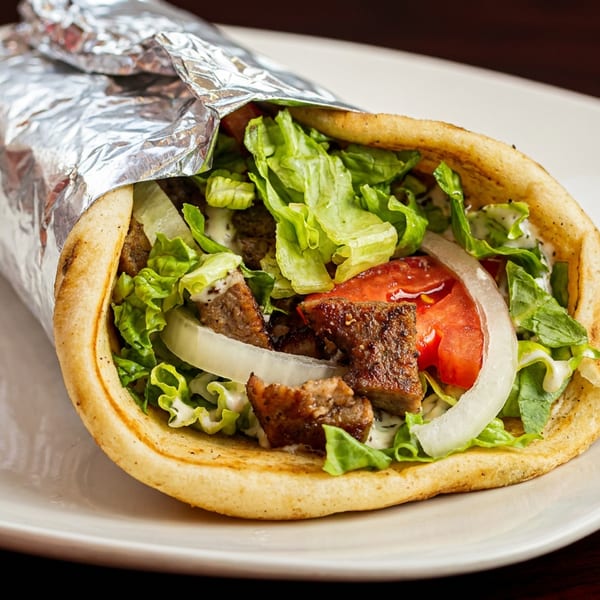 A gyro with meat, lettuce, tomato, onion, and sauce, partially wrapped in foil on a white plate.